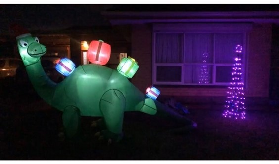 A brightly lit large dinosaur stands in front of a house. To the right is a glowing Christmas Tree 